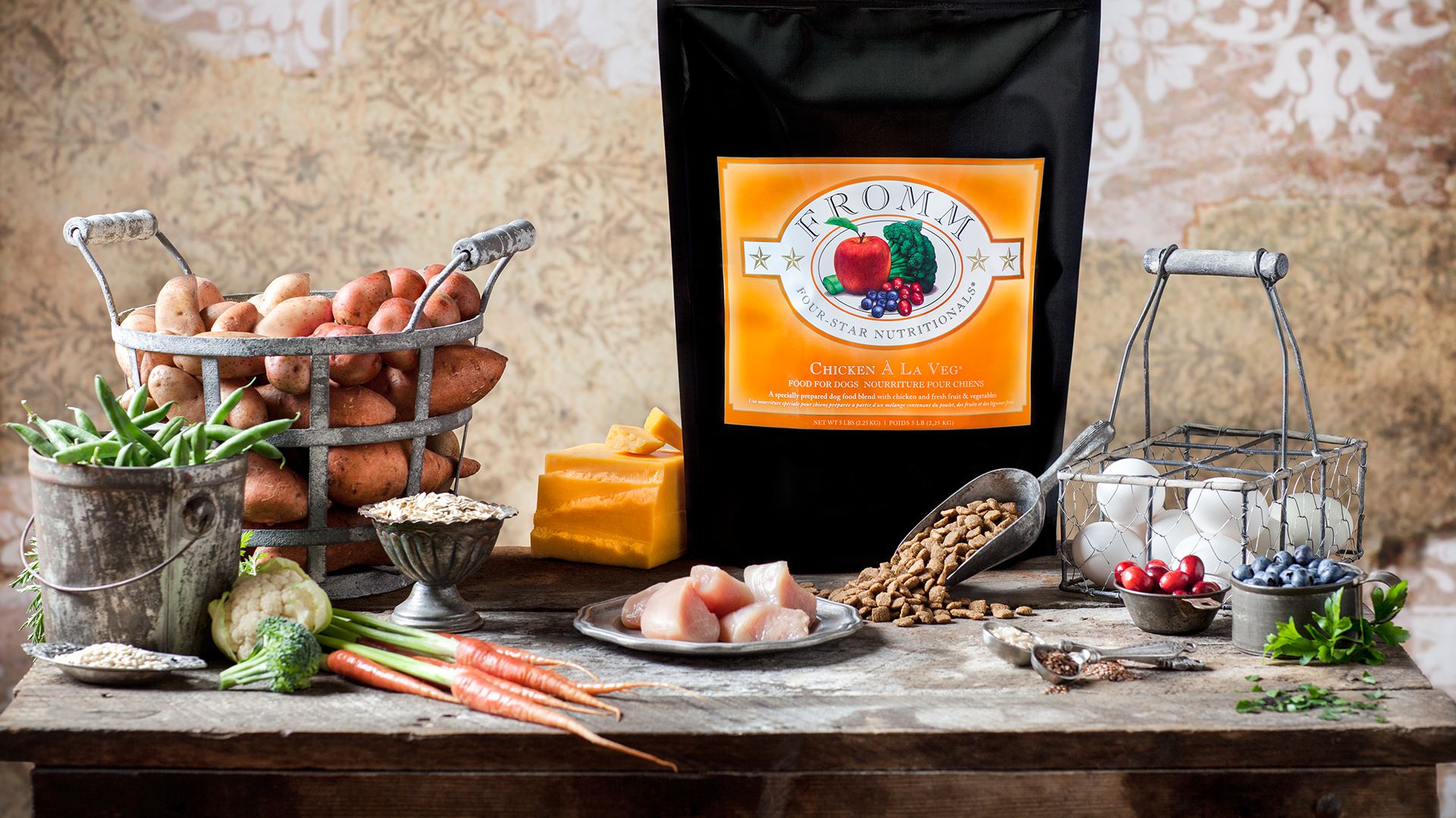 An image of the FROMM Chicken a la Veg bag of dog food resting on a harvest table surrounded by fresh vegetables, chicken and eggs.